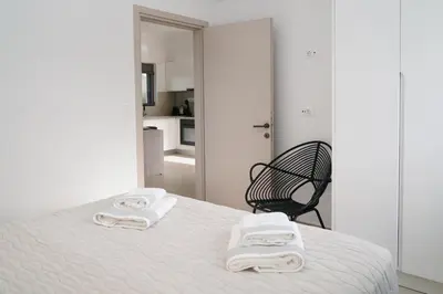 a white bedroom with a bed and a black chair at Niki Luxury Suites Pool Resort 200m From Beach in Chorafakia