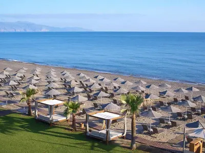 a beach with white umbrellas and palm trees and the ocean at Minoa Palace Resort in Plataniás