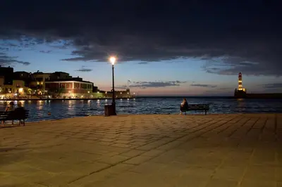 a woman sitting on a bench near the water at night at Emmanouil’s House in Chania Town