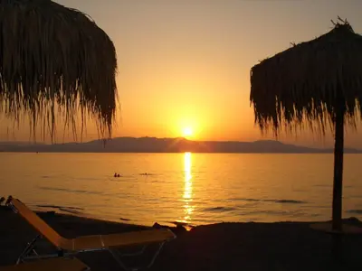 a sunset on the beach with two chairs and an umbrella at Raph's Studios-Apts By The Sea in Agia Marina Nea Kydonias